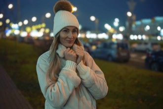 A woman wearing a cozy coat and hat stands outside on a cool evening. The city lights twinkle in