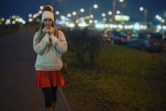 A woman walks on a city sidewalk at night, wearing a warm white jacket and a beanie. The street is