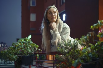 A woman with long hair leans on a railing surrounded by lush plants, reflecting on a lively city
