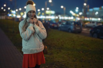 A woman stands outside on a cool evening, wearing a warm jacket and beanie. She smiles softly while