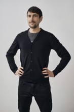 A man poses confidently with his hands on his hips, wearing a black cardigan over a gray shirt. He