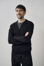 A man is posing confidently with arms crossed, wearing a black cardigan over a gray shirt. He