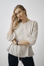 A pensive woman is standing with one hand touching her hair while wearing a light, textured blouse.