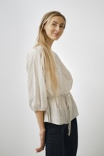 A blonde woman model stands playfully in a light-colored blouse with bell sleeves. She wears dark