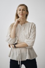A woman stands in a relaxed pose with one hand on her chin, wearing a light-colored, casual blouse.