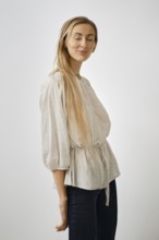 A woman stands in a plain background, wearing a light-colored blouse with a waist tie. Her long