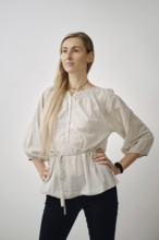 A woman stands confidently in a light beige blouse with long sleeves, paired with dark jeans. Her