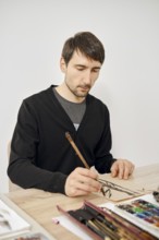 A man carefully draws on a sheet of paper in a bright studio. He uses a brush and appears deep in