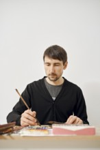 A focused artist is working diligently on a paper in a clean, bright studio. He uses a brush to add