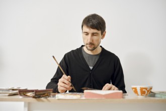 A man is concentrating on painting in a minimalist studio, surrounded by art supplies and colorful