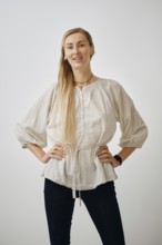 A woman stands confidently in a plain studio, wearing a light, textured blouse and dark pants. She