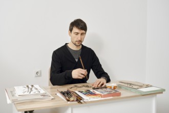 An artist sits at a wooden table, focused on painting with watercolors. The studio is bright and