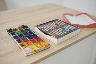 A wooden table displays two sets of art supplies. On the left, a vibrant watercolor palette with