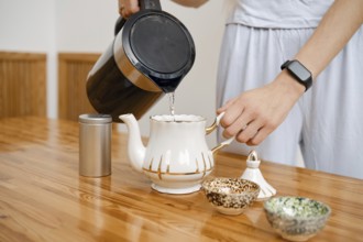A person pours hot water from a kettle into a delicate teapot. On the wooden table, small bowls are