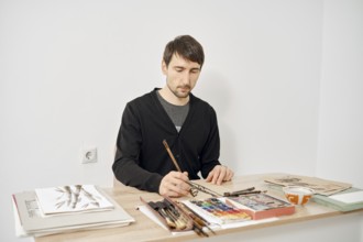 A focused artist is drawing at a wooden table in a bright studio. Various art supplies, including