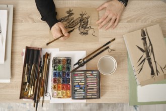 A person is painting with watercolors and sketching with fine brushes on brown paper. Art supplies