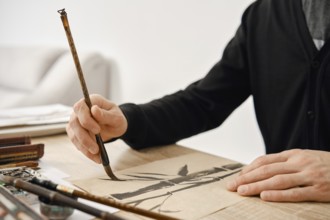 An artist is focused on creating a detailed brush painting on paper. The scene shows natural light