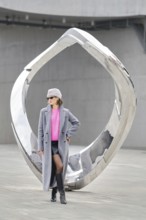 A woman stands confidently in urban attire against a shiny modern sculpture. The scene showcases