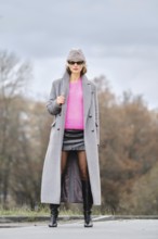 A fashionable woman in a vibrant pink sweater, a sleek black skirt, and a long gray coat standing