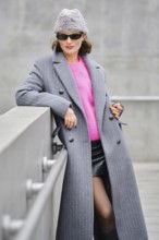 A woman stands confidently on an urban street, dressed in a long gray coat over a vibrant pink