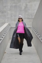Model walks confidently in urban fashion featuring bright pink sweater and stylish coat