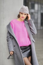 A woman stands casually against a concrete pillar in an urban setting, wearing a bright pink