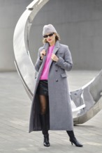 A woman in a long gray coat and pink sweater posing near a large modern sculpture. She wears black