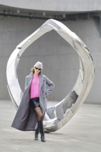 A woman wearing a vibrant pink sweater and gray coat stands next to a shiny modern sculpture. The