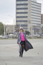 A stylish woman walks along a city sidewalk, wearing a vibrant pink sweater, leather skirt and a