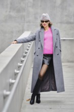 A stylish playful woman poses on a contemporary walkway, wearing a long gray coat, pink sweater,