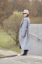 A fashionable woman poses on steps wearing a long grey coat, high-heeled boots, and a stylish