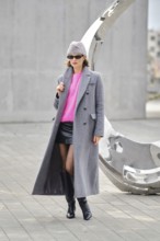 In an urban setting, a woman posing in a long gray coat over a pink sweater and a black leather