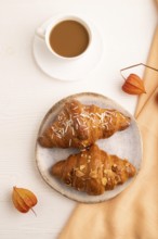 Croissant on blue plate on white wooden background and orange linen textile, cup of coffee, top