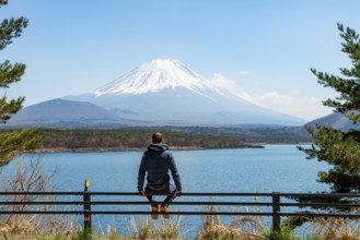 Young man sitting on railings next to a road and looking across the lake to Mt Fuji volcano, Motosu
