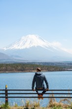 Young man sitting on railings next to a road and looking across the lake to Mt Fuji volcano, Motosu