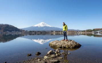 Young man standing on a rock in water, volcano Mt. Fuji is reflected in Lake Kawaguchi, Yamanashi