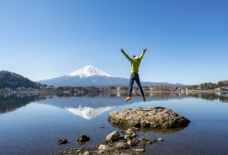 Young man standing on a rock in water, jumping into the air, volcano Mt. Fuji is reflected in Lake