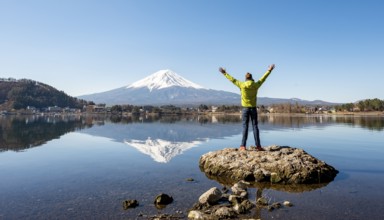 Young man stands on a rock in water, stretches his arms in the air, volcano Mt. Fuji is reflected