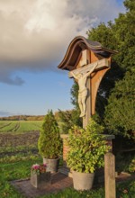 Wooden cross with Jesus figure against the backdrop of a rural landscape in sunlight, Münsterland,