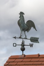 Metal weather vane with rooster on a tiled roof against cloudy sky, Baden-Württemberg, Germany