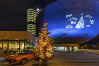 Illuminated Christmas tree with red balls, next to a historic BMW vehicle, in front of the BMW