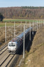 A passenger train travels through an autumnal landscape with fields and forests in the background,