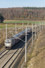 A passenger train travels on a railway line through an autumnal landscape with fields and forests,