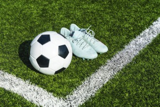 Soccer ball and football cleats sitting on a green artificial grass pitch near the white boundary