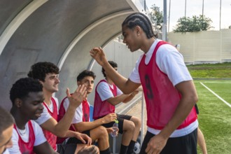 Soccer players in red bibs sitting on the bench and high fiving in the dugout, sharing a moment of