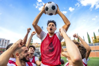 Young men soccer teammates in red bibs celebrate victory, raising a ball overhead with joyful