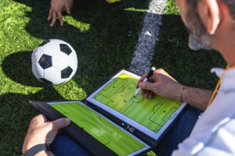 Soccer coach sketching tactical game plan on a clipboard beside a ball on artificial grass near a