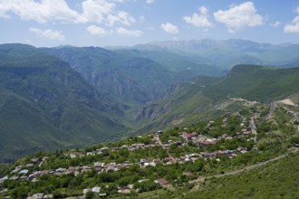 A small village is situated in a green valley surrounded by forested mountains under a cloudy sky,