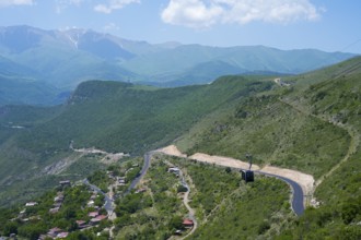 A winding road runs through green hills and passes a small village, with views of mountains, Tatev