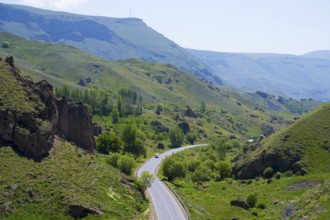 A winding road leads through a green valley with hills and trees under a blue sky, landscape